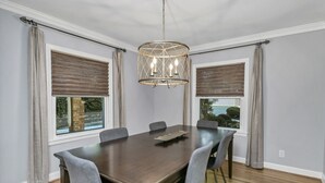 A dining room with a large wooden table, six grey upholstered chairs, a chandelier, and two windows with brown blinds. - Forest Lake Retreat | Backyard Sanctuary & Hot Tub (Columbia)
