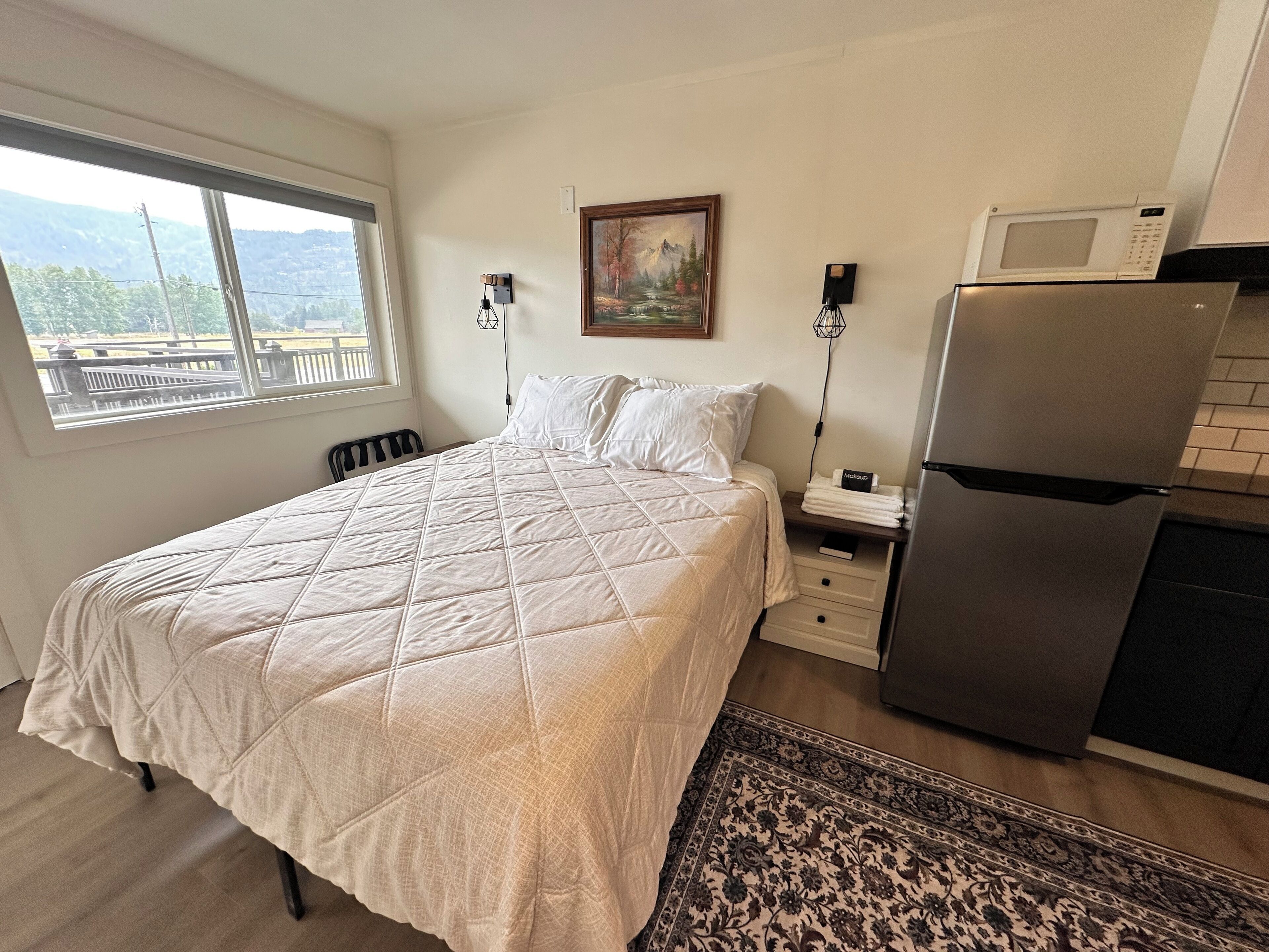 Quarto com cama, mesa de cabeceira, geladeira, janela com vista e um quadro na parede.