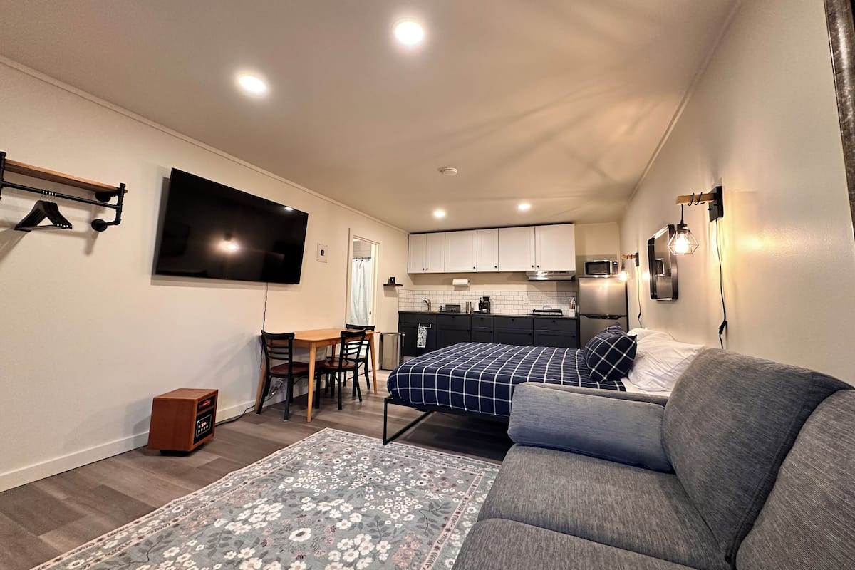 A compact living space with a sofa, a TV mounted on the wall, a small kitchen area, and a dining table.