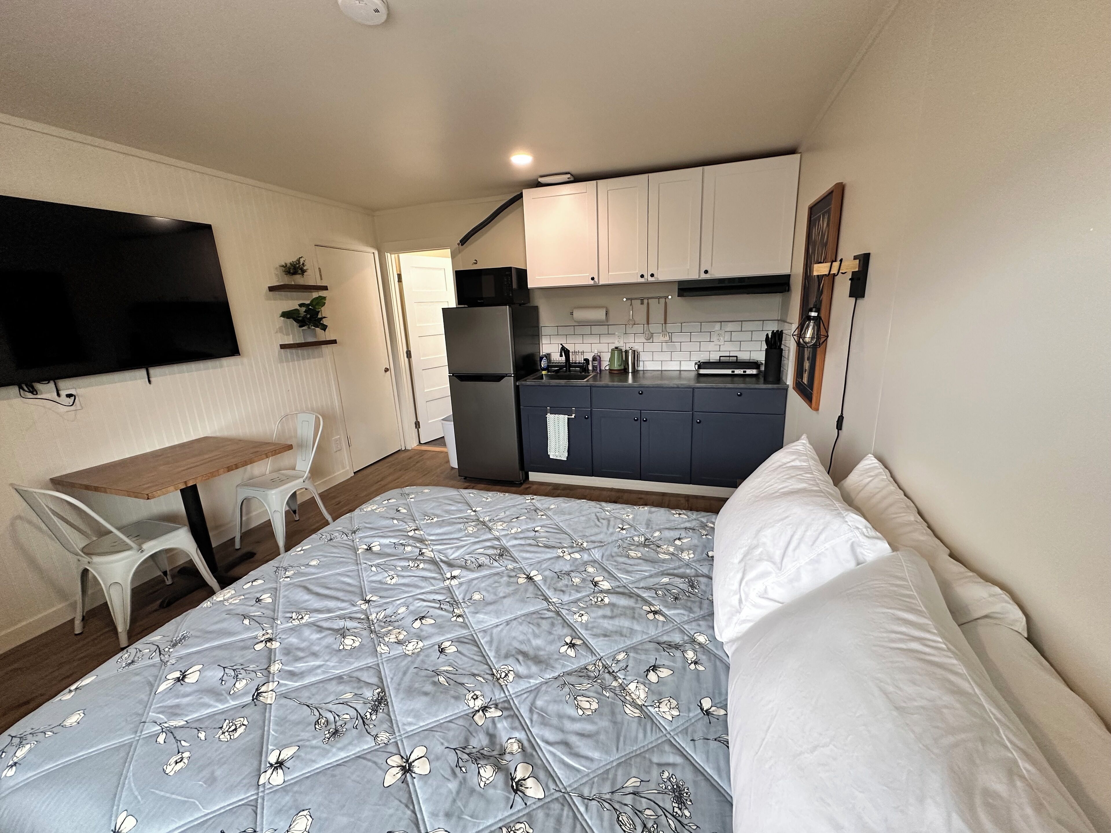 Una habitación compacta con cama, una pequeña mesa de comedor, una zona de cocina y un televisor montado en la pared.