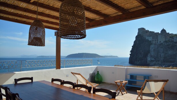 A terrace with a wooden table and chairs, a hanging wicker light fixture, and a view of a coastal cliff and sea. - In an old fishing village apartment with sea view terrace (Ischia)
