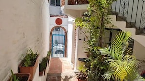 A narrow, sunlit corridor with potted plants on either side, leading to a blue door with a decorative arch.