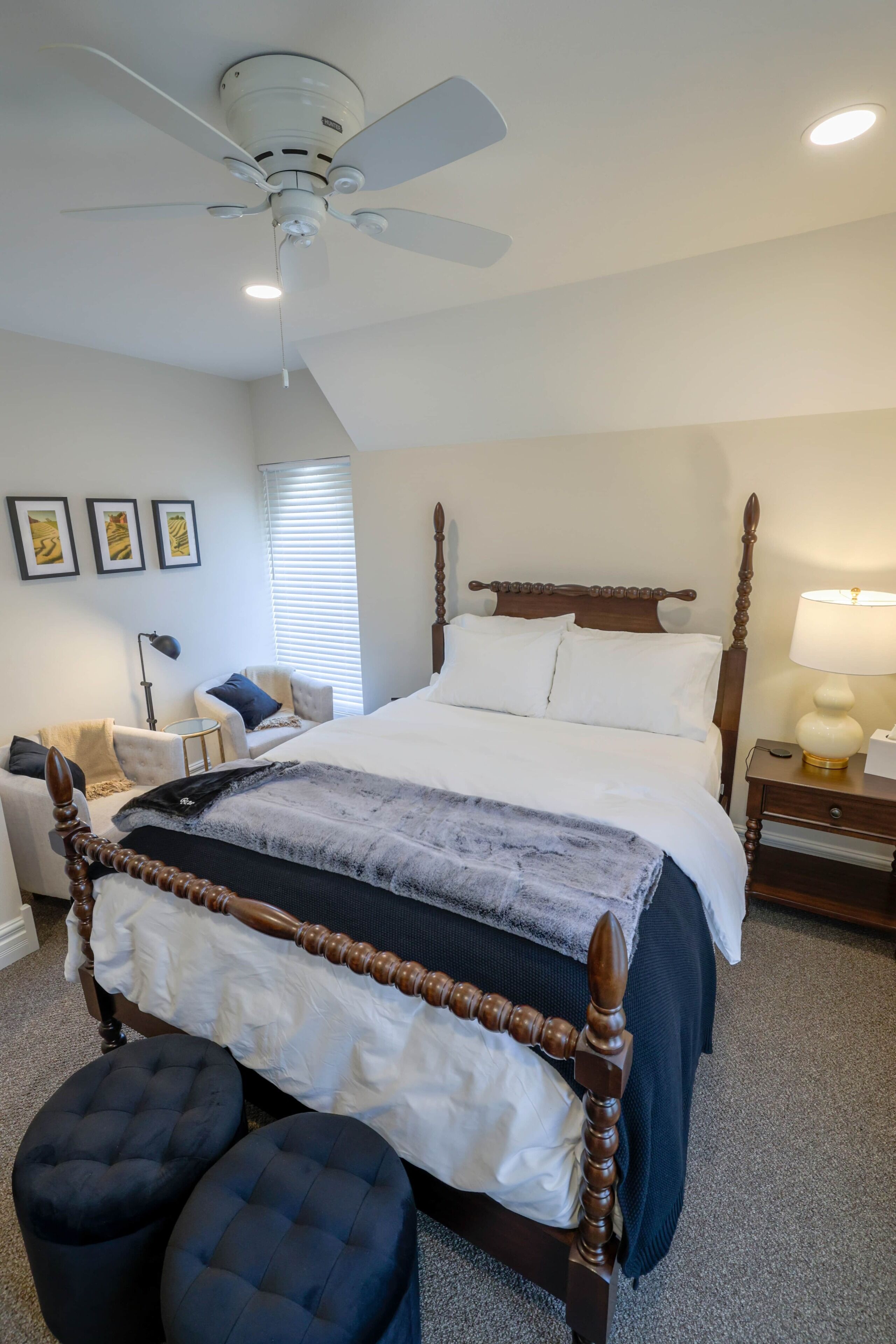 A neatly arranged bedroom with a wooden four-poster bed, a ceiling fan, a bedside lamp, and a small table with a lamp.