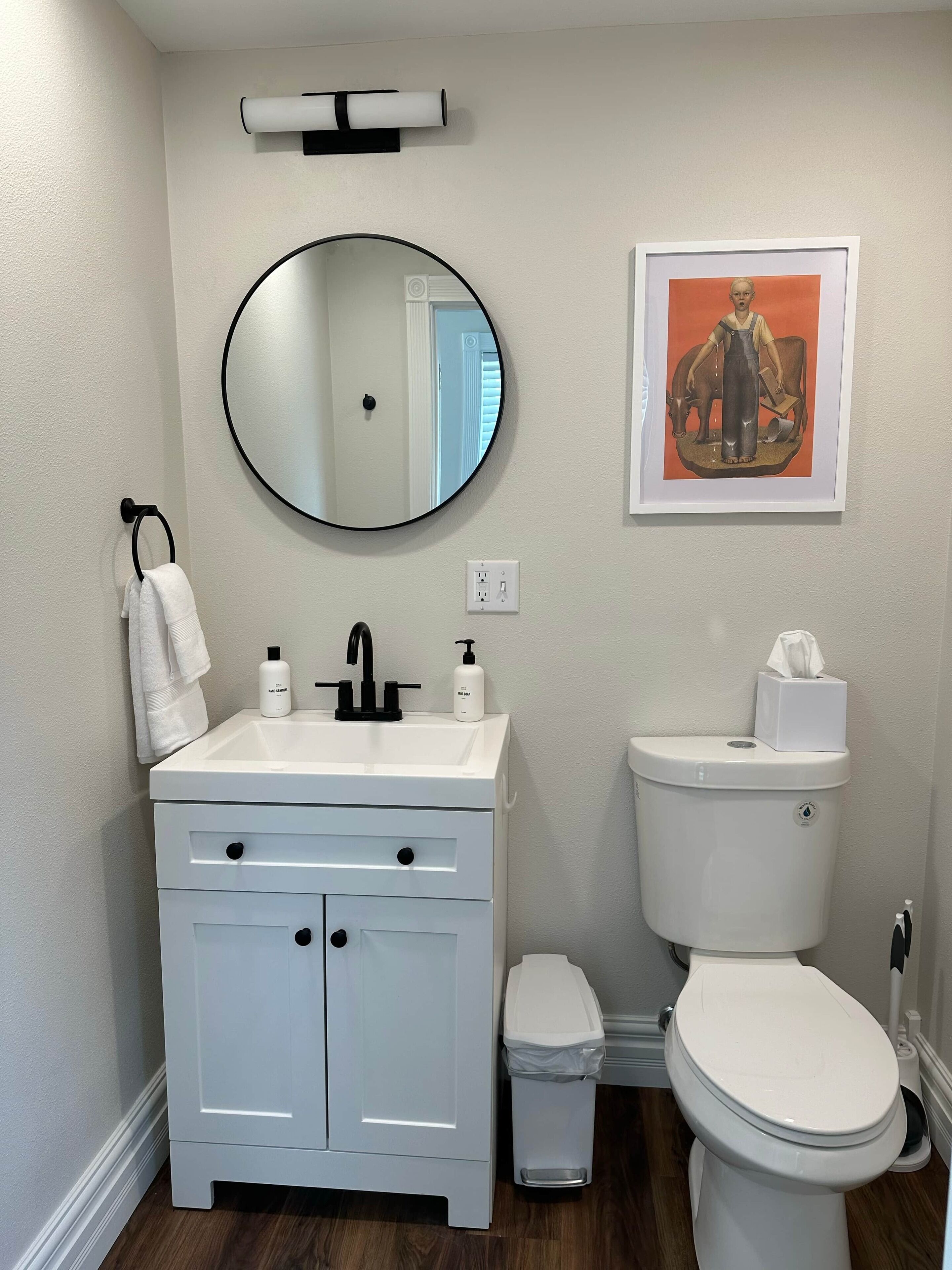 Une salle de bain avec un lavabo blanc, un miroir et des toilettes. Un tableau encadré est accroché au mur, et une poubelle se trouve à côté des toilettes.