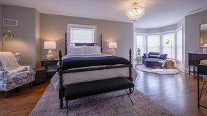 A bedroom with a large bed, two bedside tables with lamps, a chair, a sofa, and a large window with blinds. - Riggs House Restored 1908 Home (Maquoketa)