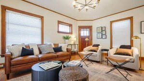 A living room with a brown leather sofa, two white armchairs, a coffee table, and a woven ottoman. There are framed pictures on the wall and a chandelier above. - Riggs House Restored 1908 Home (Maquoketa)