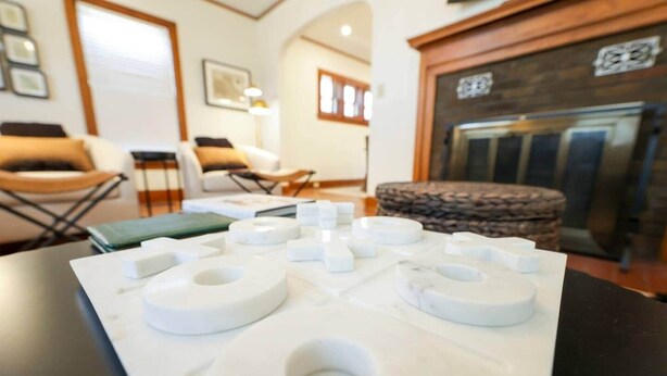 A room with a fireplace, a table with white circular objects, and a chair. - Riggs House Restored 1908 Home (Maquoketa)