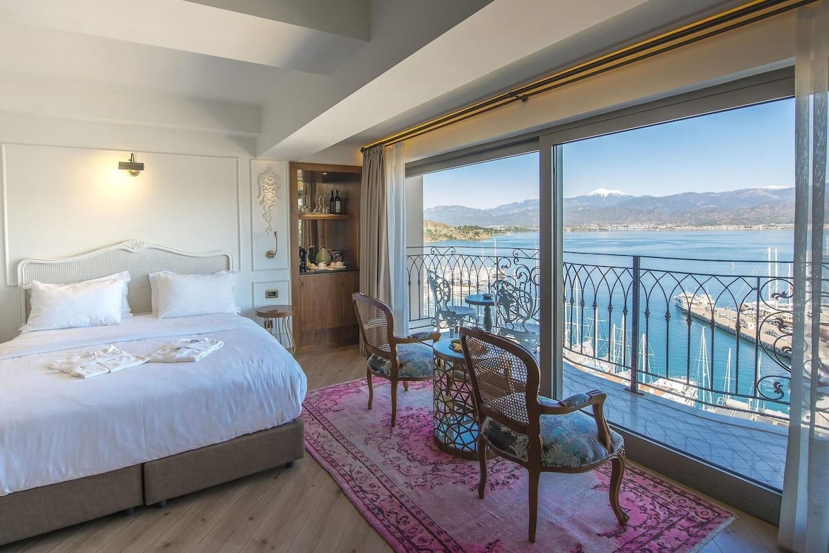 A bedroom with a large bed, a small table, and a chair, a balcony with a view of a body of water and mountains, and a wooden shelf with bottles.