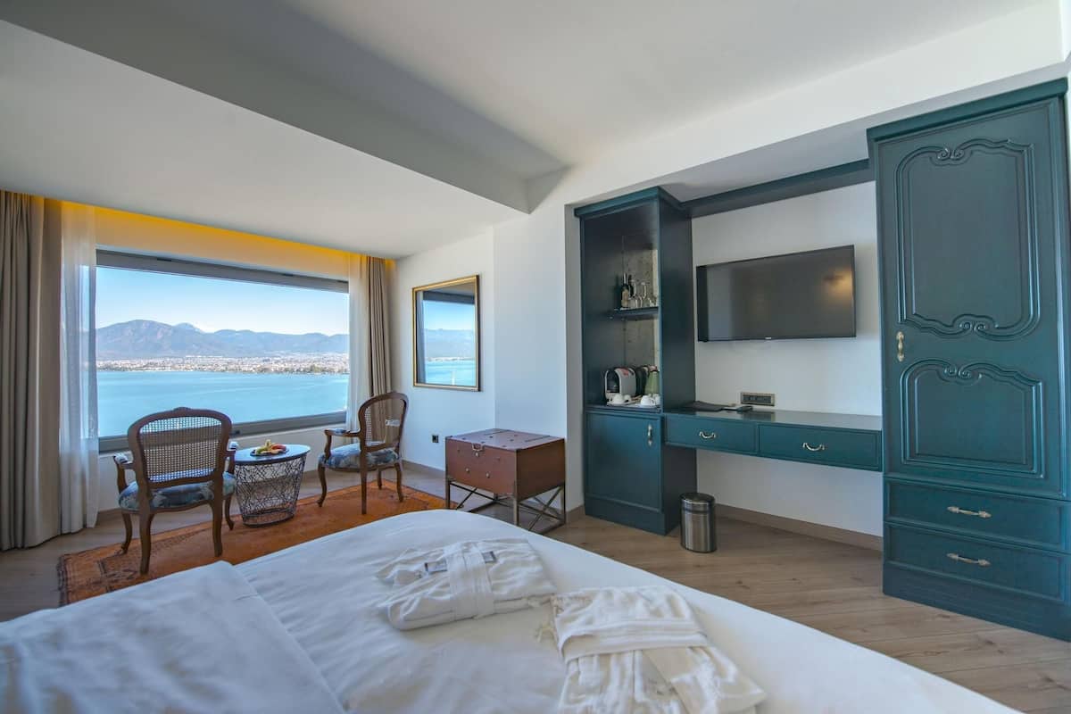 A hotel room with a bed, a desk, a chair, a TV, and a view of the sea.