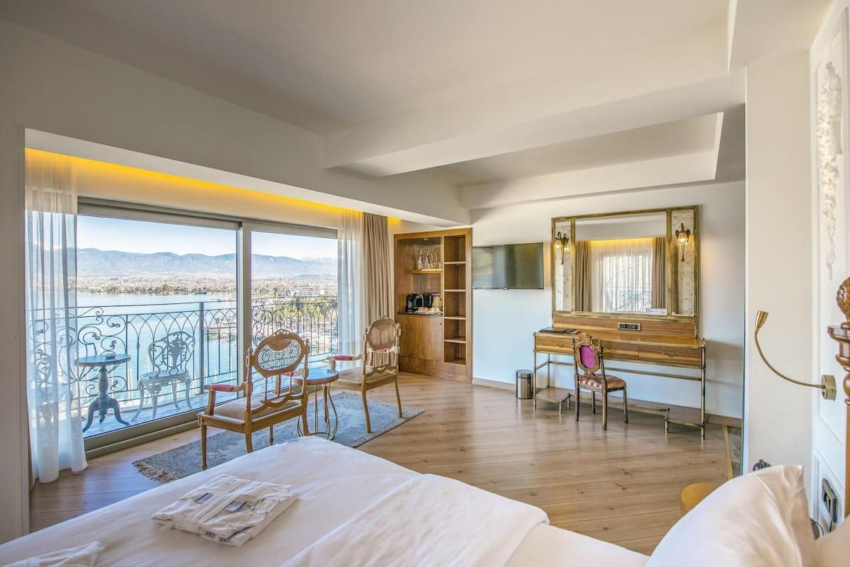 A hotel room with a large bed, a desk, a chair, a wardrobe, and a balcony with a view of the mountains.