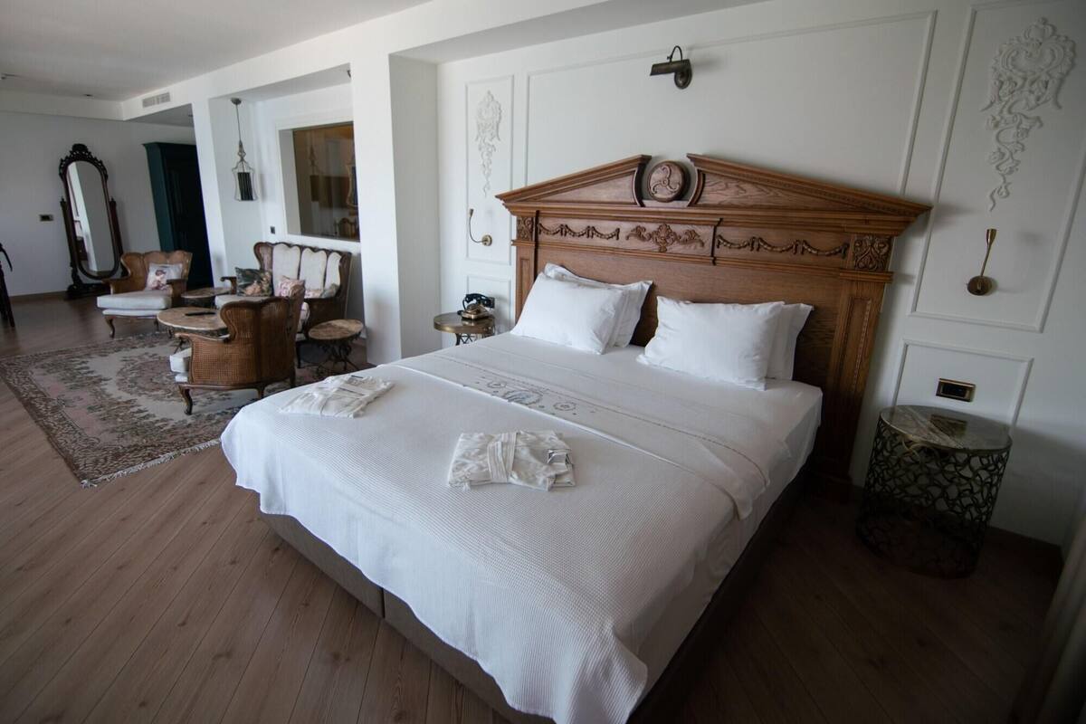 A hotel room with a large bed, a wooden headboard, a nightstand, a chair, a mirror, and a decorative wall panel.