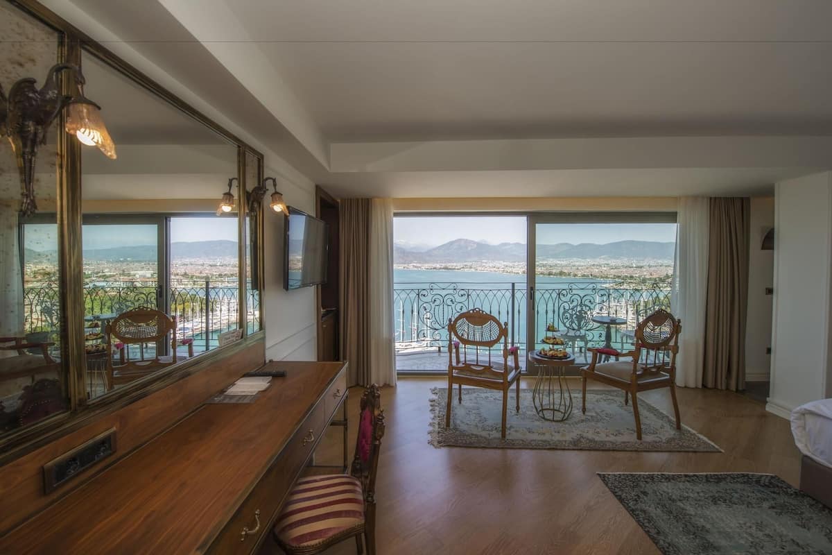 A room with a wooden desk, a large mirror, a television, and a view of a city and mountains through large windows.
