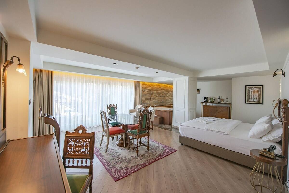 A hotel room with a bed, a desk, a chair, a small table, a bathtub, and a window with curtains.