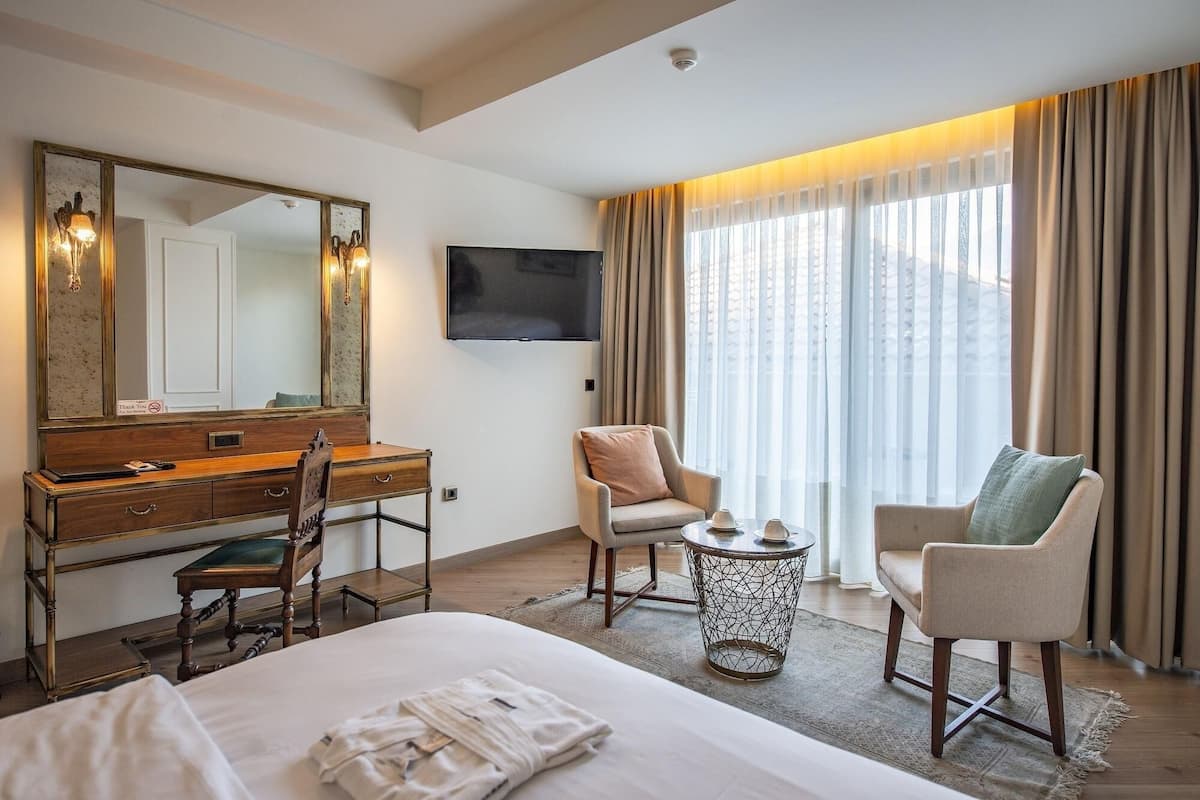 A hotel room with a bed, a desk with a chair, a round side table, a large mirror, and a television on the wall.
