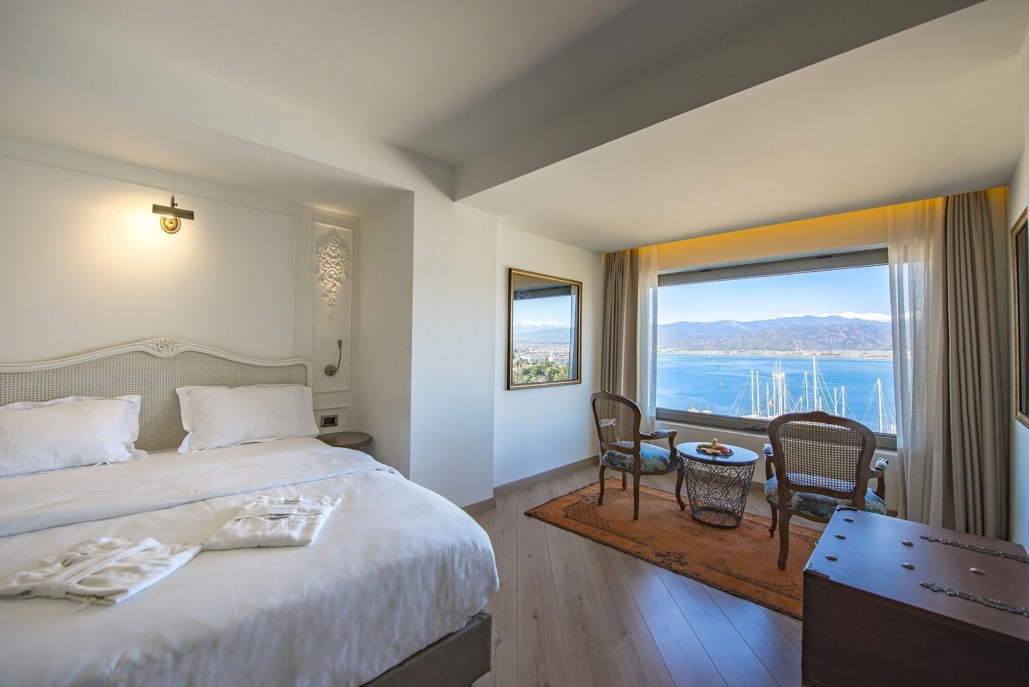 A hotel room with a bed, a small table with chairs, a view of the sea, and a window with curtains.