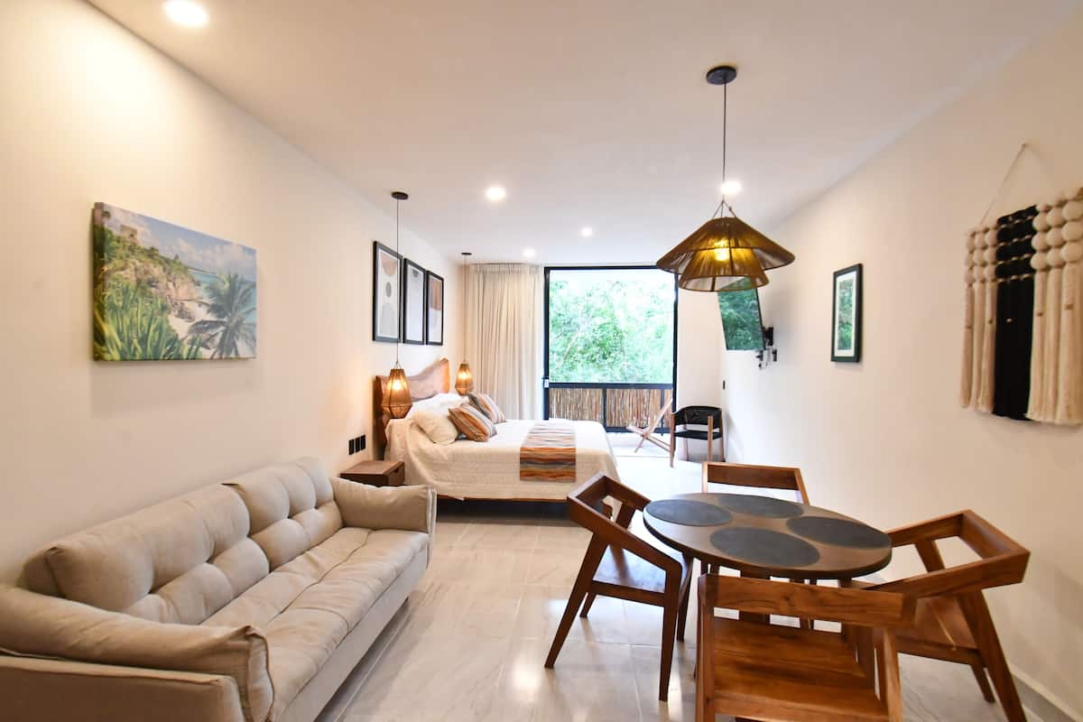 A modern bedroom with a large bed, bedside tables, a wooden dining table with chairs, a sofa, and a large window with a view of greenery.