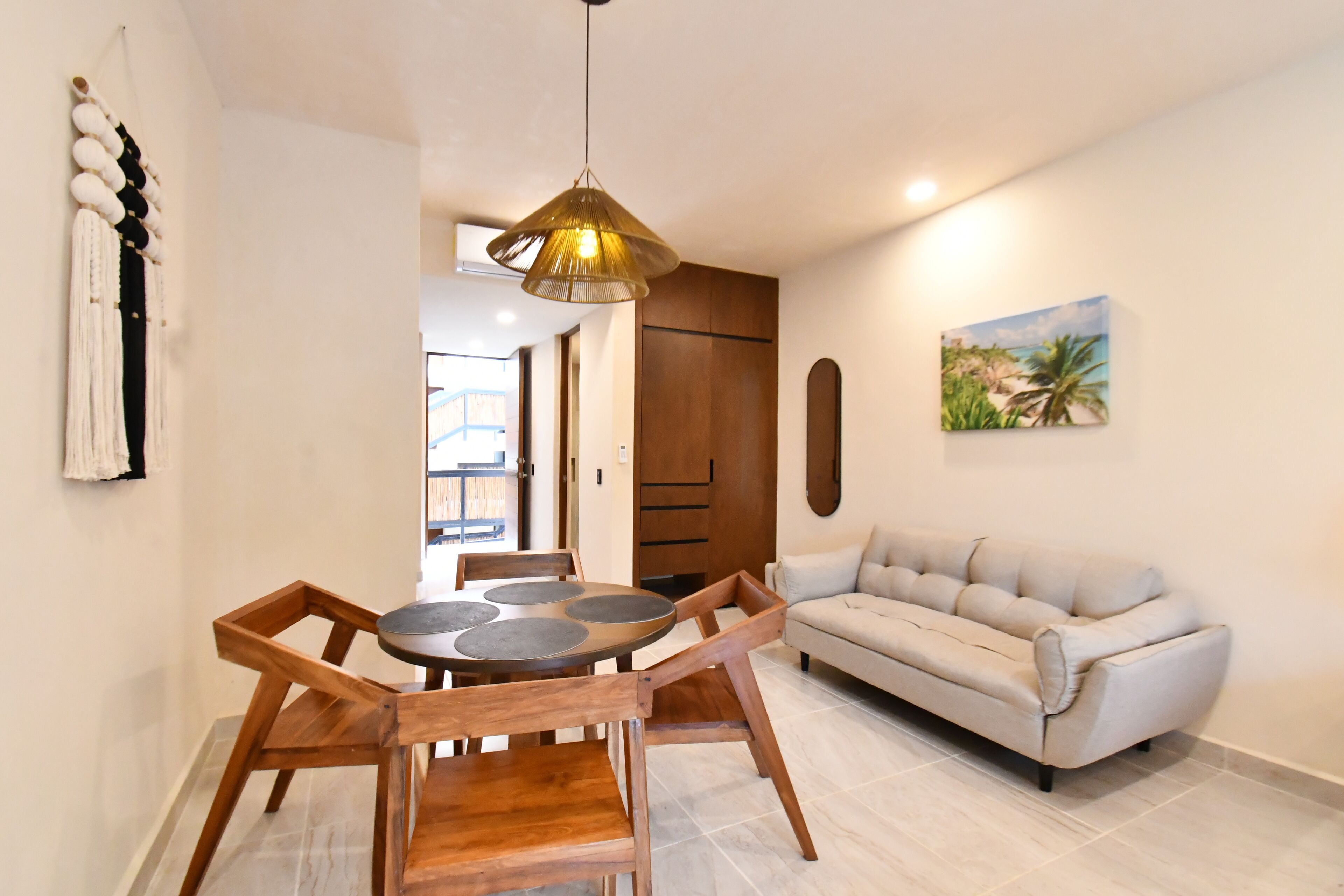 A dining area with a round wooden table and chairs, a beige sofa, a hanging light fixture, and a painting on the wall.