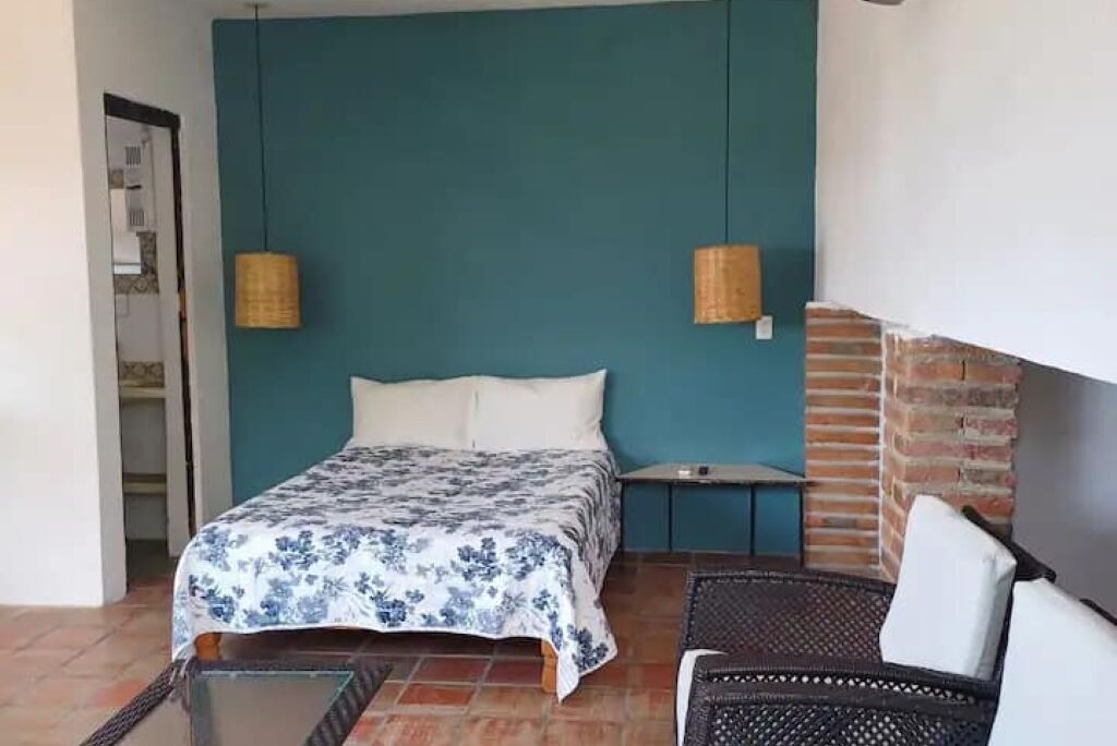 Un dormitorio con cama, mesita de noche, silla de mimbre y lámpara colgante.