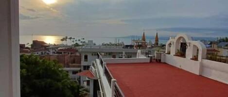 Eine Dachterrasse mit rotem Boden, weißen Geländern und Blick auf Stadt und Meer bei Sonnenuntergang.