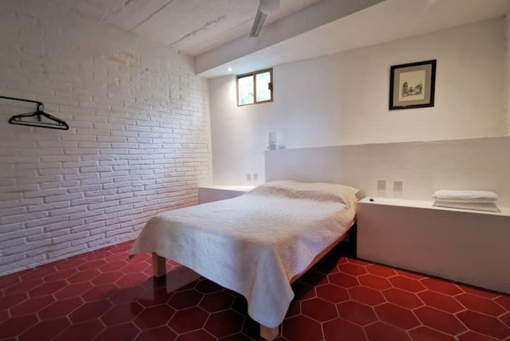 Een slaapkamer met een bakstenen muur, een bed met een witte spreidek, een rode hexagonale vloer, een wit nachtkastje en een klein raam.