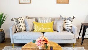 Un canapé gris clair agrémenté de plusieurs coussins à motifs, d’un coussin décoratif avec l’inscription « Bienvenue » et d’une petite table sur laquelle est disposé un arrangement floral.