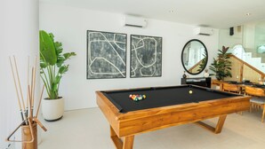 A modern living room with a pool table, two abstract paintings, a potted plant, and a staircase leading to an upper level. - Modern 3br Villa with Rooftop Patio - Pool Table (South Kuta)