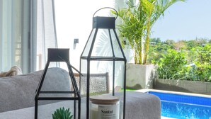 A modern living room with a glass table displaying a candle and a small plant. A large window offers a view of a swimming pool and palm trees. - Modern 3br Villa with Rooftop Patio - Pool Table (South Kuta)