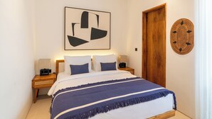 A modern bedroom with a large bed, wooden headboard, a wall-mounted abstract art piece, a round woven wall hanging, and a wooden door. - Modern 3br Villa with Rooftop Patio - Pool Table (South Kuta)