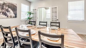 A dining room with a wooden table set for six, surrounded by chairs. There is a large window with blinds, a door with glass panels, and a chandelier.