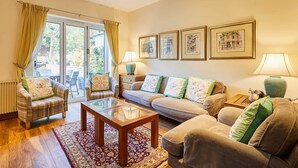A living room with a sofa, armchair, coffee table, and a large window with curtains. There are framed pictures on the wall and a lamp on a side table. - Host Stay The Station Cottage (Rowlands Gill)