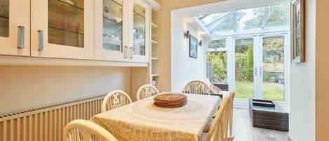 A dining area with a table set for four, surrounded by white chairs. There is a glass cabinet above the table with dishes and glassware. A large window provides a view of the garden outside.