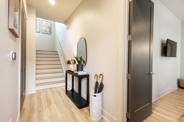 Un couloir avec un escalier, une console noire surmontée d’un miroir, une poubelle blanche et une porte sombre.