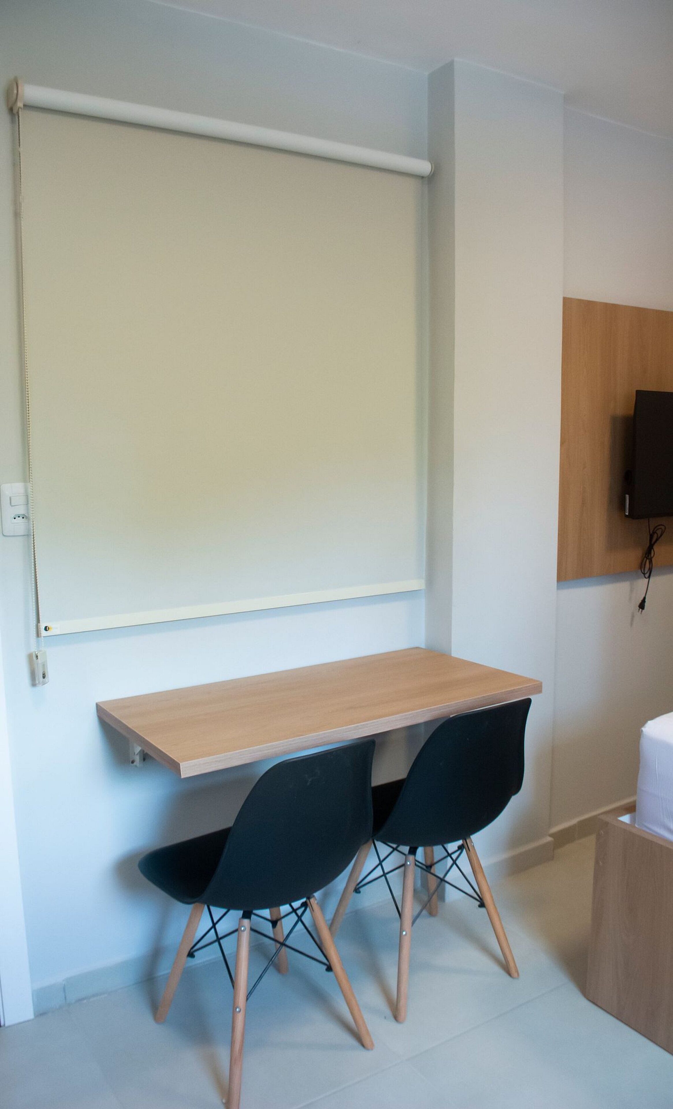 A room with a wooden desk, two black chairs, a projector screen, a mounted TV, and a power outlet.