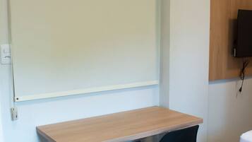 A room with a wooden desk, two black chairs, a projector screen, a mounted TV, and a power outlet.