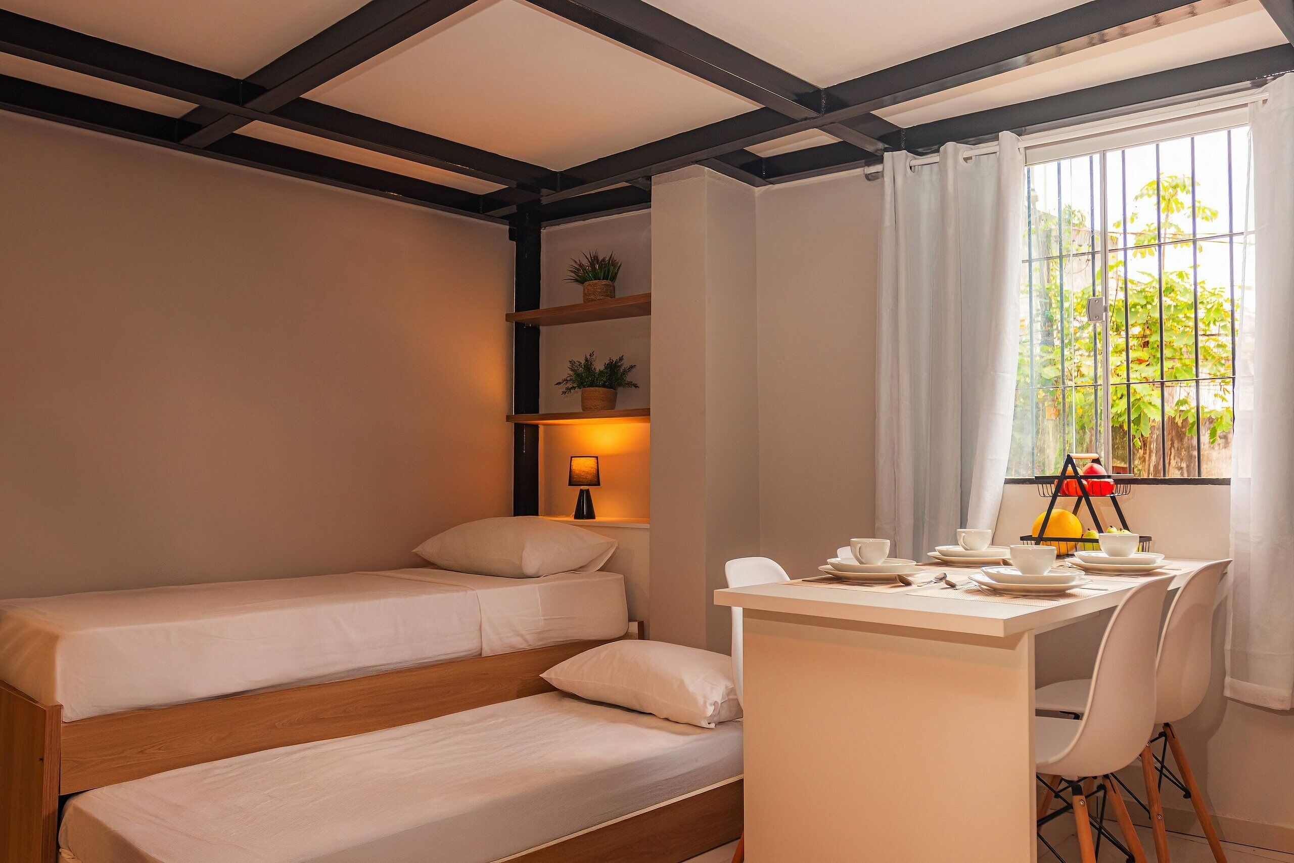 Ein Hotelzimmer mit zwei Einzelbetten, einem Schreibtisch mit Stühlen, einem kleinen Tisch mit Lampe und einem Fenster mit Vorhängen.