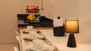 Une table à manger bien dressée, avec de l’argenterie blanche, des tasses et un set de table. On aperçoit, à l’arrière-plan, une lampe noire et un panier de fruits.