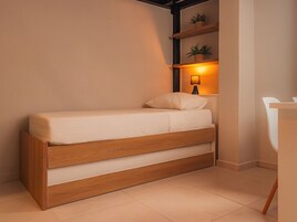Una habitación pequeña con cama individual, estructura de madera, mesita de noche, lámpara y estante con plantas.