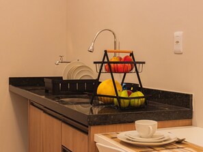 Una cocina con encimera negra, un fregadero, una cesta de frutas y una taza sobre un platillo.