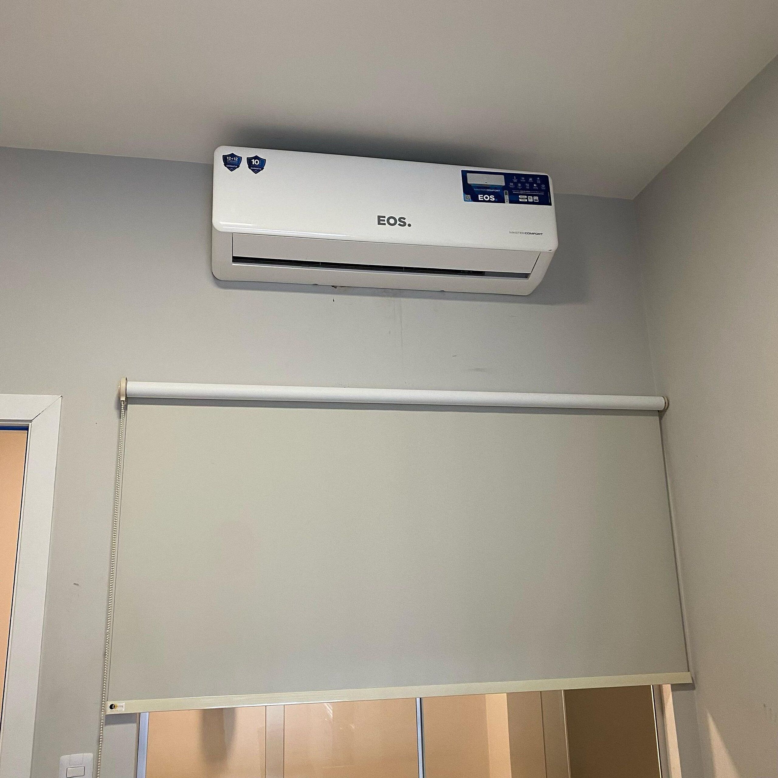 A white air conditioning unit mounted on a wall above a window with a beige blind.
