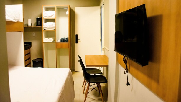 Une chambre d’hôtel compacte comprenant un lit, un bureau, une chaise, un téléviseur fixé au mur et une armoire avec des étagères.