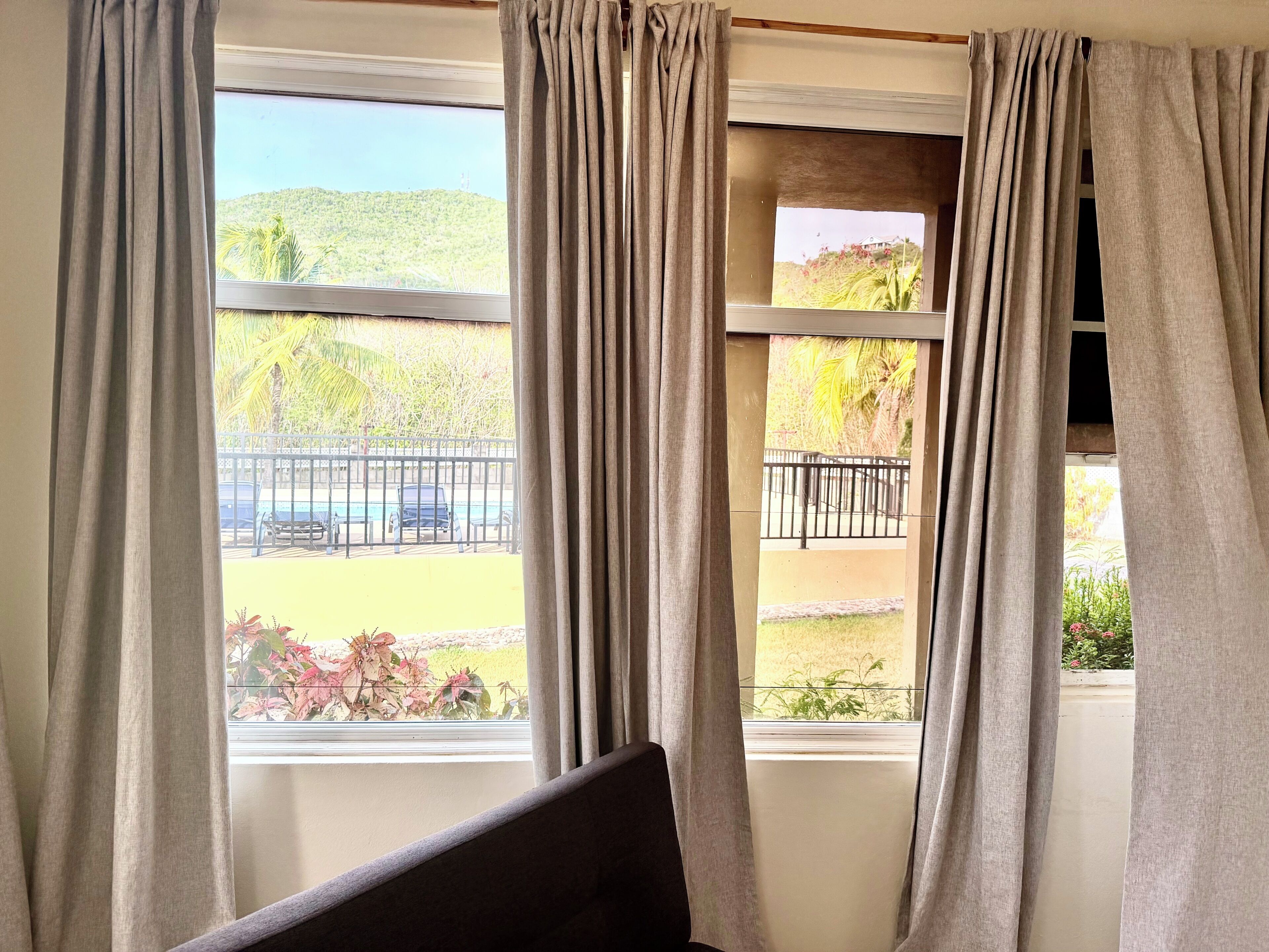 Habitación con dos ventanas, cada una con cortinas, y vista a un estacionamiento con valla y vegetación.