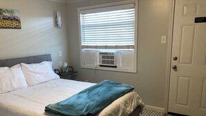 A neatly made bed with white linens and a blue blanket, a bedside table with a tissue box, a wall-mounted air conditioner, and a door with a 'No Smoking' sign. - All-inclusive studio apartment with utilities included-Unit 5 (Waterville)
