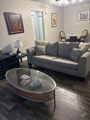 A living room with a grey sofa, a glass coffee table, a black side table, and a lamp. There are framed pictures on the walls and a door with blinds. - Cozy Sandy Springs Haven (Sandy Springs)