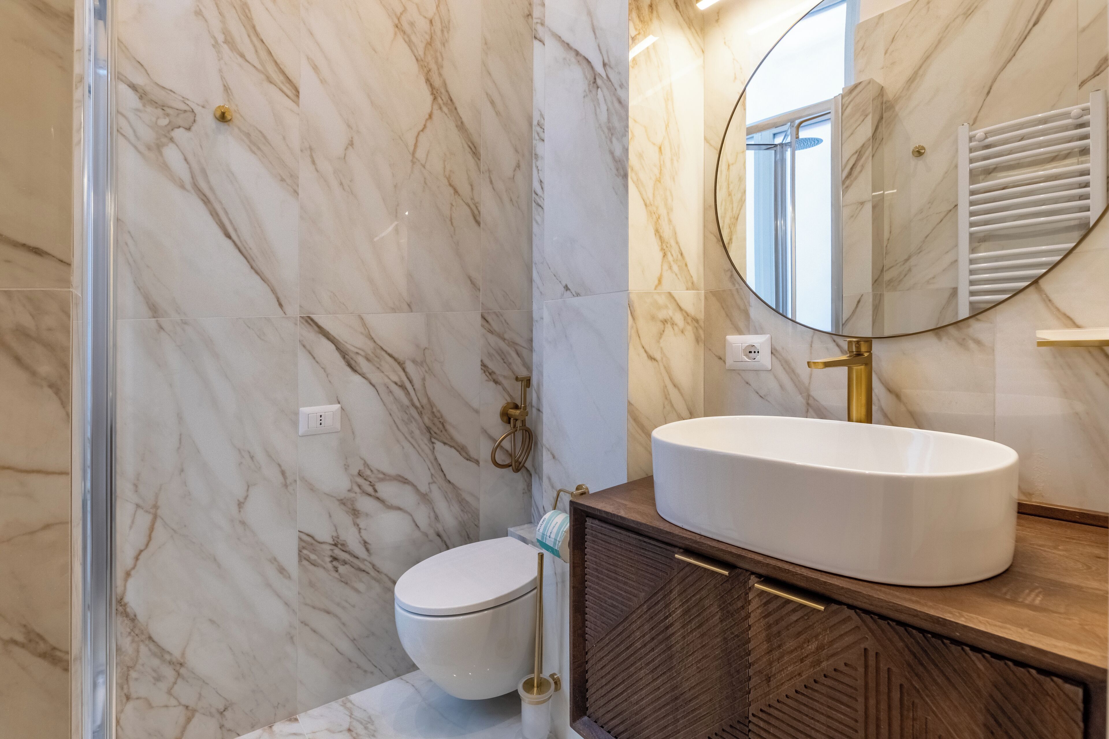 A bathroom with a marble wall, a white toilet, a round sink, and a wooden cabinet.