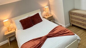 Ein ordentlich bezogenes Bett mit weißer und brauner Bettwäsche, zwei Nachttischlampen und eine Holzkommode in einem Zimmer mit Holzboden und schrägverlaufender Decke.
