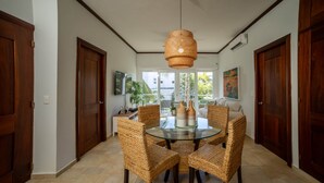 A dining area with a round glass table surrounded by wicker chairs, a woven pendant light, and a view of the outdoors through large windows. - Bright & Modern 2BR/2BA Apartment Comfort & Style In cabarete (Cabarete)