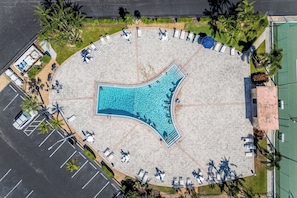 Une vue aérienne d’une piscine entourée de chaises longues et de parasols, avec des palmiers et un court de tennis en arrière-plan.