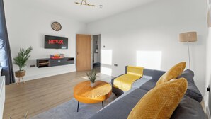 Ett modernt vardagsrum med en grå soffa, gula kuddar, ett runt, orange soffbord, en tv som är monterad på väggen och en trädörr som leder till ett annat rum.