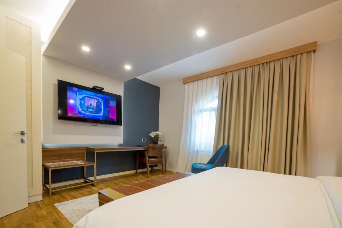 A hotel room with a bed, a desk, a chair, a TV mounted on the wall, and a window with curtains.
