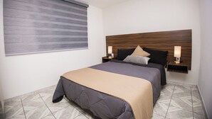 A modern bedroom with a large bed, a wooden headboard, two bedside lamps, and a window with blinds. - House with large kitchen, 1 block from third ring road (Aguascalientes)